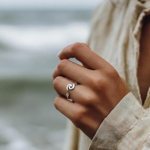 May include: A silver ring with a wave design. The ring is worn on a finger, with the wave shape curving across the top. The ring is made of a shiny, reflective metal. The background is blurred, with a hint of a beach scene.