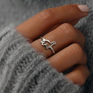 May include: A silver-colored ring featuring a stylized outline of a hand gesture. The ring is worn on a finger, and the design is simple and modern. The ring is a piece of jewelry.