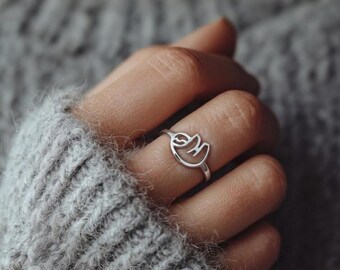 May include: A silver ring featuring a sloth design. The ring is a minimalist style, with the sloth's outline forming the ring's centerpiece. The ring is worn on a finger, with a gray sweater in the background.