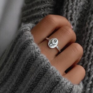 Puede incluir: Anillo de plata con una cara ovalada que muestra un dise&ntilde;o grabado. El anillo est&aacute; en un dedo, sobre un su&eacute;ter de punto gris. El dise&ntilde;o del anillo es un p&aacute;jaro o pluma estilizado. Una pieza de joyer&iacute;a.