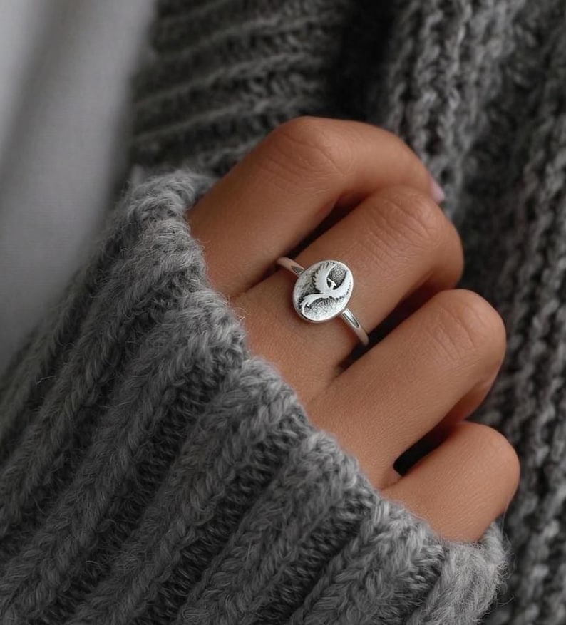 Puede incluir: Anillo de plata con una cara ovalada que muestra un dise&ntilde;o grabado. El anillo est&aacute; en un dedo, sobre un su&eacute;ter de punto gris. El dise&ntilde;o del anillo es un p&aacute;jaro o pluma estilizado. Una pieza de joyer&iacute;a.