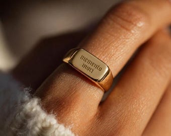 May include: A gold signet ring with the inscription "memento mori" in a classic font. The ring has a rectangular face and a polished finish. The ring is worn on a finger.