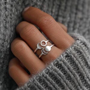 May include: Two silver rings with celestial designs. One ring features a circle, and the other has a crescent moon and a full moon. The rings are displayed on a gray background.