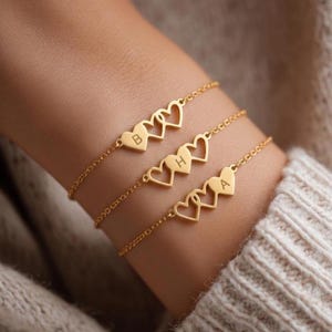 May include: Three gold-colored bracelets, each featuring a delicate chain and heart-shaped charms. Each charm has a letter engraved on it. The bracelets are worn on a wrist, with the letters "B", "H", and "A" visible.