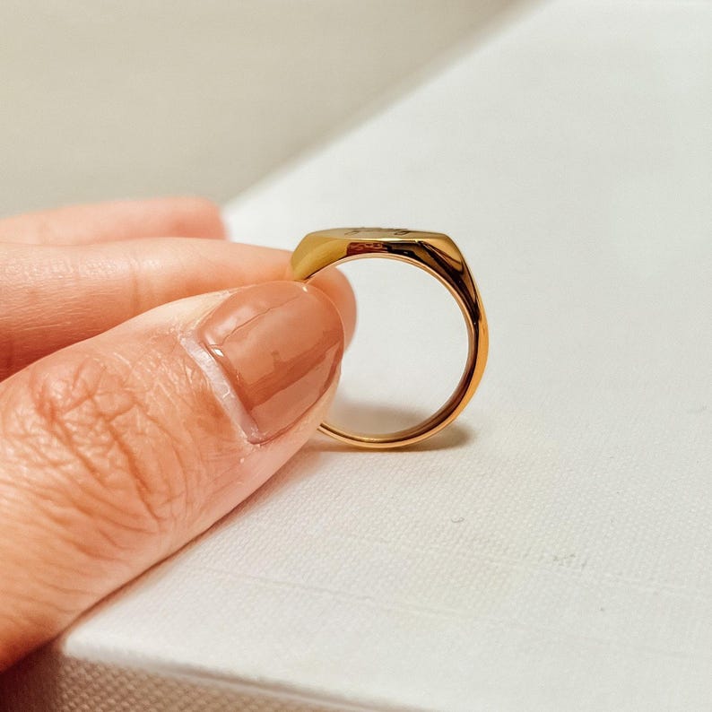 K&ouml;nnte beinhalten: Ein goldfarbener Siegelring mit rechteckiger Oberfl&auml;che. Der Ring wird hochgehalten, um seine polierte Oberfl&auml;che und sein Design zu pr&auml;sentieren. Der Ring hat ein glattes, abgerundetes Band.