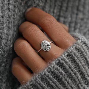 May include: A silver ring featuring an oval-shaped design with a detailed fern leaf engraving. The ring is shown on a hand, with a gray sweater in the background. The ring is a piece of jewelry.