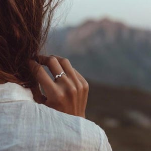 May include: A silver-colored ring with a mountain range design. The ring is worn on a finger, with a blurred mountain backdrop. The ring's design features a series of peaks and valleys, creating a minimalist and nature-inspired aesthetic.