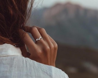 Op de afbeelding: Een zilverkleurige ring met een bergketenontwerp. De ring wordt aan een vinger gedragen, met een vage bergachtergrond. Het ontwerp van de ring heeft een reeks pieken en valleien, waardoor een minimalistische en door de natuur geïnspireerde esthetiek ontstaat.