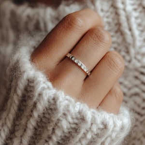 May include: A silver ring with a series of small, circular, moon-like designs. The ring is worn on a finger, with a soft, textured, light-colored sweater in the background. The ring's design is repeated around the band.