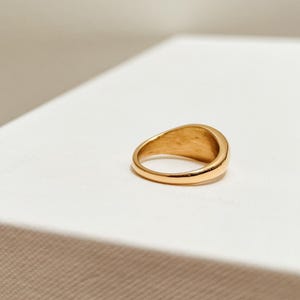 K&ouml;nnte beinhalten: Ein goldfarbener Siegelring mit glattem, abgerundetem Design. Der Ring besteht aus einem gl&auml;nzenden, reflektierenden Metall und liegt auf einer wei&szlig;en Oberfl&auml;che. Das Band des Rings hat eine gleichm&auml;&szlig;ige Breite, und die Oberseite hat eine leicht gebogene, ovale Form.
