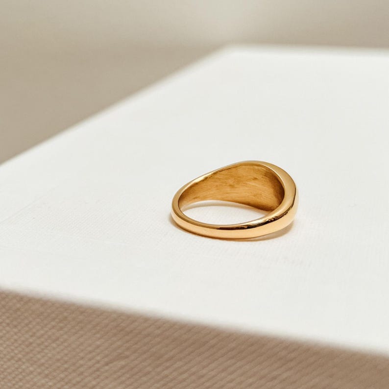 K&ouml;nnte beinhalten: Ein goldfarbener Siegelring mit glattem, abgerundetem Design. Der Ring besteht aus einem gl&auml;nzenden, reflektierenden Metall und liegt auf einer wei&szlig;en Oberfl&auml;che. Das Band des Rings hat eine gleichm&auml;&szlig;ige Breite, und die Oberseite hat eine leicht gebogene, ovale Form.