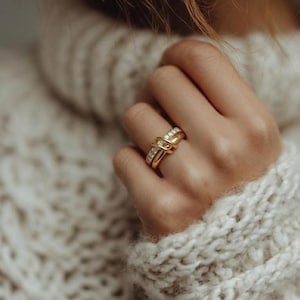 May include: A close-up of a gold ring with a knot design, adorned with small, sparkling gemstones. The ring is on a hand, set against a backdrop of a cozy, cream-colored knit sweater. The ring's intricate design and the sweater's texture create a visually appealing contrast.