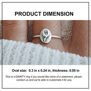 Puede incluir: Anillo de plata con un colgante ovalado con un dise&ntilde;o de f&eacute;nix. El &oacute;valo mide 0,76 cm por 0,61 cm, con un grosor de 0,13 cm. El anillo se muestra en un dedo, sobre un fondo blanco.