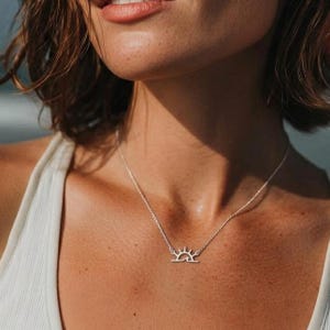 May include: A silver necklace featuring a minimalist sun design pendant. The pendant depicts a stylized sun with rays and a curved base. The necklace is worn on a person's neck, against a white top.