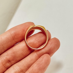 K&ouml;nnte beinhalten: Ein goldfarbener Ring mit rechteckiger Oberseite und glatter, polierter Oberfl&auml;che. Der Ring wird in einer Hand gehalten, was sein schlichtes Design hervorhebt. Das Band des Rings ist abgerundet, und die Gesamt&auml;sthetik ist minimalistisch.