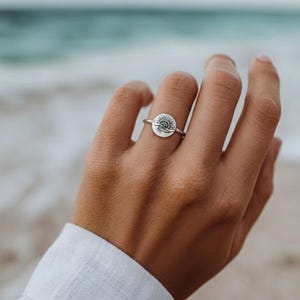 May include: A silver ring featuring a circular design with a sun and wave motif. The ring is worn on a finger, with the ocean in the background. The ring's band is thin and delicate, and the design is detailed.