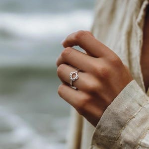 May include: A silver ring featuring a turtle design. The ring is worn on a finger, with the turtle design facing forward. The ring is made of a shiny metal and has a simple, elegant design.
