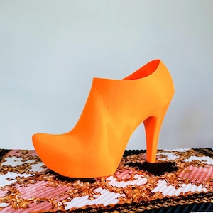 May include: A bright orange high-heeled shoe with a pointed toe. The shoe is sitting on a patterned fabric with gold and black accents.