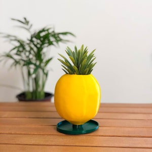 May include: A yellow pineapple-shaped planter with a green base, holding a small green plant.