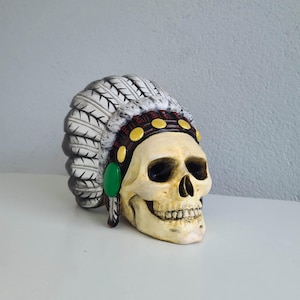 Könnte beinhalten: Ein weißer und schwarzer Schädel, der eine rot-schwarze Kopfbedeckung mit goldenen Akzenten und einer grünen Feder trägt. Der Schädel hat eine weiße und schwarze Federkopfbedeckung mit einem rot-schwarzen Band mit goldenen Akzenten.