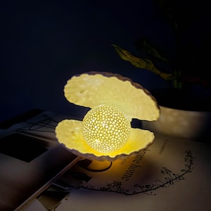 May include: A white ceramic night light shaped like an open clam shell with a glowing pearl inside. The pearl has a textured surface and is illuminated from within.