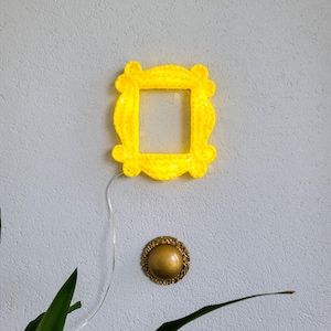 Op de afbeelding: Een heldergele, verlichte fotolijst met een vierkante opening, gemonteerd op een lichtgrijze muur. Onder het frame bevindt zich een goudkleurige, ronde deurklopper. Een dunne, zilveren draad loopt van het frame.