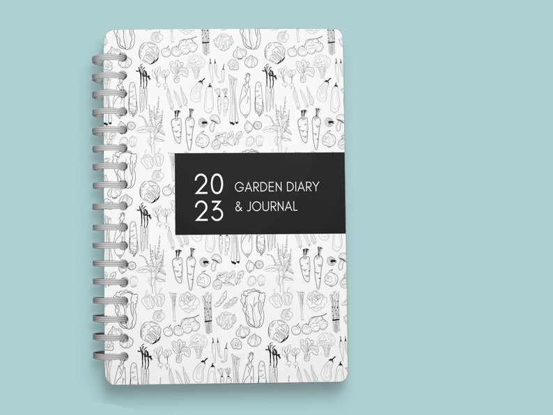 Pu&ograve; includere: Un diario bianco a spirale con un'illustrazione in bianco e nero di verdure sulla copertina. Il diario &egrave; intitolato "2023 Garden Diary & Journal".