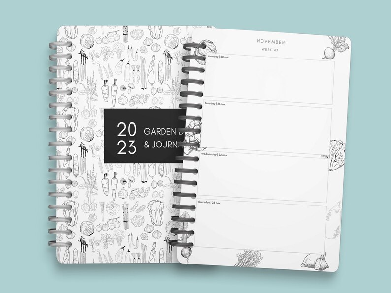 Pu&ograve; includere: Un diario a spirale bianco e nero con una copertina che presenta un motivo di verdure disegnate a mano. La copertina ha anche un'etichetta nera con il testo "2023 GARDEN & JOURNAL". Le pagine interne sono vuote e presentano un calendario per novembre, settimana 47.