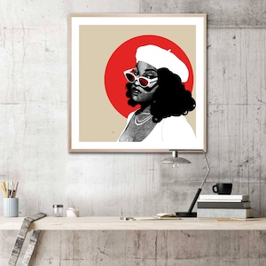 May include: A framed art print featuring a stylized portrait of a person wearing a white beret and sunglasses with red accents. The portrait is set against a tan background with a large red circle. The artwork is in black, white, and red.