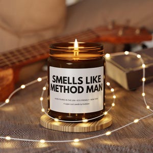 May include: A lit brown glass candle jar with the text "SMELLS LIKE METHOD MAN" on a white label. The candle sits on a small wooden coaster, with a string of lights and a guitar in the background.