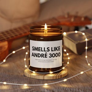Op de afbeelding: Een brandende bruine glazen potkaars met een wit etiket met de tekst "SMELLS LIKE ANDRÉ 3000". De kaars staat op een kleine houten onderzetter, met een slinger van feeërieke lichtjes eromheen. Een gitaar en een boek staan op de achtergrond.