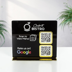 Può includere: Un espositore nero e oro con codice QR per Orbit Bistro. L'espositore presenta il logo del ristorante, le parole "Scan to View Menu!" e "Rate us on! Google." Vengono visualizzati due codici QR, insieme al nome del ristorante.