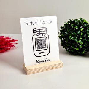 May include: A white acrylic sign with the text "Virtual Tip Jar" and "Thank You". The sign features a black line drawing of a mason jar with a QR code and the word "venmo". The sign is set in a wooden base.