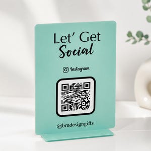 May include: A teal acrylic sign with the text "Let' Get Social" and a QR code for Instagram. The sign also includes the Instagram logo and the handle @brndesigngifts. The sign is designed to stand upright.