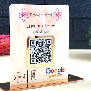 May include: A decorative sign for Flower Hotel with a QR code for reviews. The sign has a gold border and a pink floral design at the top. The sign reads "Leave Us A Review" and "Thank You". The Google logo is at the bottom.