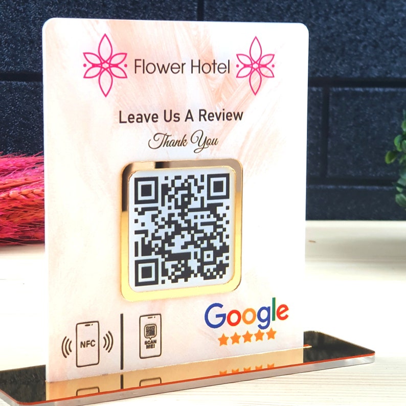 May include: A decorative sign for Flower Hotel with a QR code for reviews. The sign has a gold border and a pink floral design at the top. The sign reads "Leave Us A Review" and "Thank You". The Google logo is at the bottom.