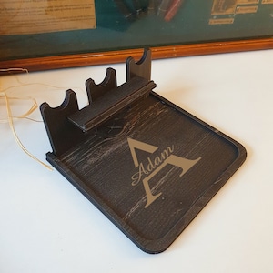 May include: A dark brown wooden phone stand with a built-in tray and three slots for charging cables. The stand has the name "Adam" engraved on it.