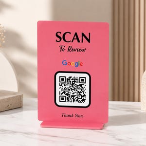 May include: A pink QR code sign with the words "SCAN To Review" and "Thank You!" printed in black. The Google logo is also visible. The sign is on a white marble surface.