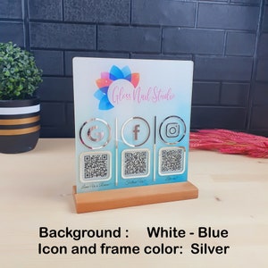 May include: A clear acrylic sign for Gloss Nail Studio, featuring a white and blue background with silver icons for Google, Facebook, and Instagram. Includes QR codes and a wooden base. The sign reads "Leave Us a Review!", "Follow Us!", and "Like us!"