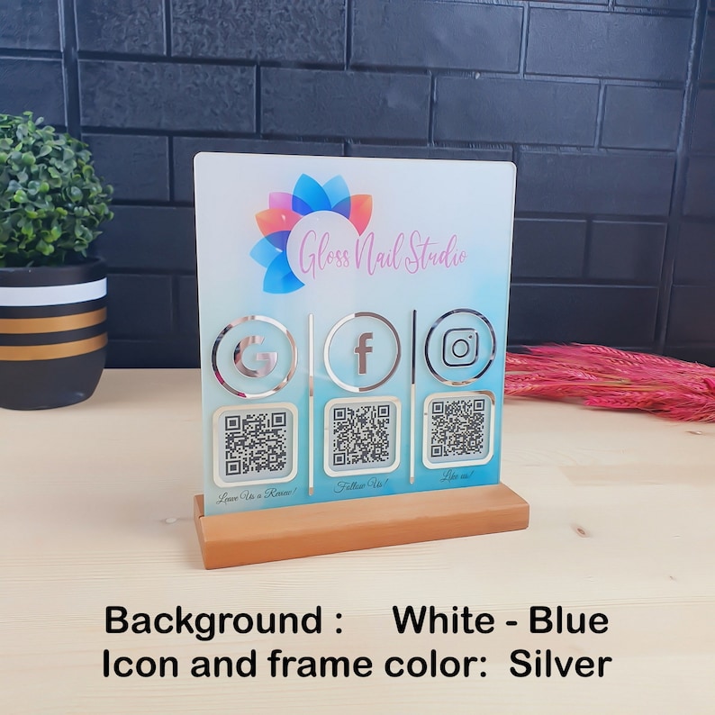 May include: A clear acrylic sign for Gloss Nail Studio, featuring a white and blue background with silver icons for Google, Facebook, and Instagram. Includes QR codes and a wooden base. The sign reads "Leave Us a Review!", "Follow Us!", and "Like us!"