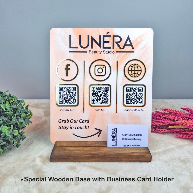 May include: A wooden business card holder with a sign for Lun&eacute;ra Beauty Studio. The sign has social media icons, QR codes, and the text "Follow Us!", "Like Us!", and "Connect With Us!" The business card displays contact information.