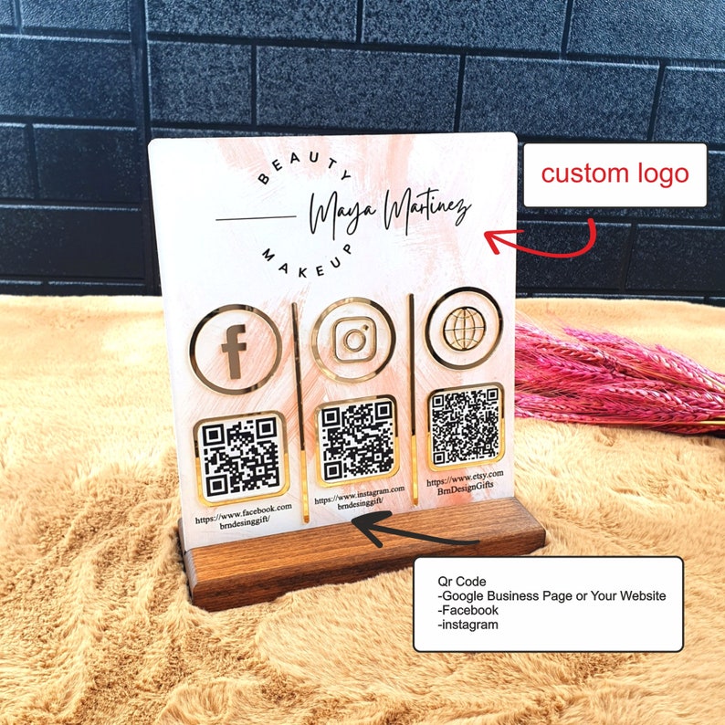 May include: A custom business sign with a wooden base. The sign features a beauty makeup theme with social media icons and QR codes for Facebook, Instagram, and a website. The sign includes the name "Maya Martinez".
