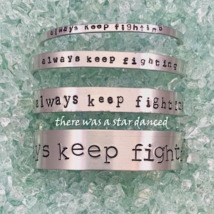 Może przedstawiać: Cztery srebrne bransoletki z wygrawerowanym napisem "always keep fighting". Dolna bransoletka ma dodatkowy tekst "there was a star danced".