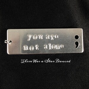 May include: A silver metal keychain with the text "you are not alone" and a semicolon engraved on it. The keychain is on a black fabric background. The text "There Was a Star Danced" is written below the keychain.
