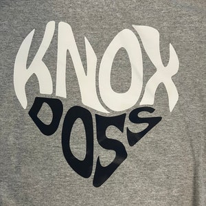 May include: A gray t-shirt with a white and black heart design. The heart design features the words "Knox" and "Boss" in a stylized font.