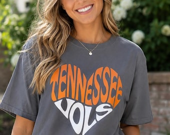 Könnte beinhalten: Ein anthrazitfarbenes Kurzarm-T-Shirt mit einem herzförmigen Aufdruck auf der Brust. Die obere Hälfte des Herzens ist orange mit dem Text TENNESSEE in fetter Schrift, die untere Hälfte ist weiß mit dem Text VOLS in fetter Schrift.