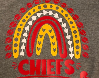 Puede incluir: Una sudadera gris con un diseño de arcoíris rojo, amarillo y blanco. El arcoíris está formado por tres arcos con corazones blancos y balones de fútbol alrededor del exterior. La palabra "CHIEFS" está escrita en rojo debajo del arcoíris.