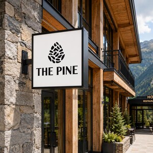 May include: A square, white, wall-mounted sign with a black frame featuring a black pinecone graphic and the text THE PINE in black lettering. The sign is attached to a stone wall of a building with wooden accents and mountain views.