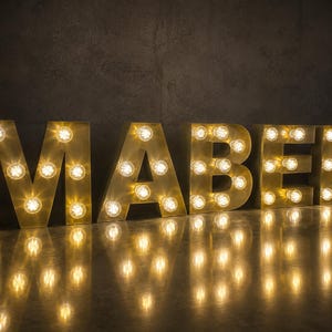 May include: A set of five large, metallic marquee letters spelling the name MABEL. Each letter features a gold-toned finish and is illuminated by multiple round, warm-toned incandescent light bulbs arranged along the front face of the letters.