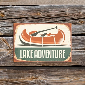 Peut inclure: Panneau rectangulaire au design vintage représentant un canoë avec une pagaie, dans les tons orange, marron et gris. Les mots "LAKE ADVENTURE" sont imprimés en blanc sur une bannière verte. Le panneau est posé sur un fond en bois vieilli.